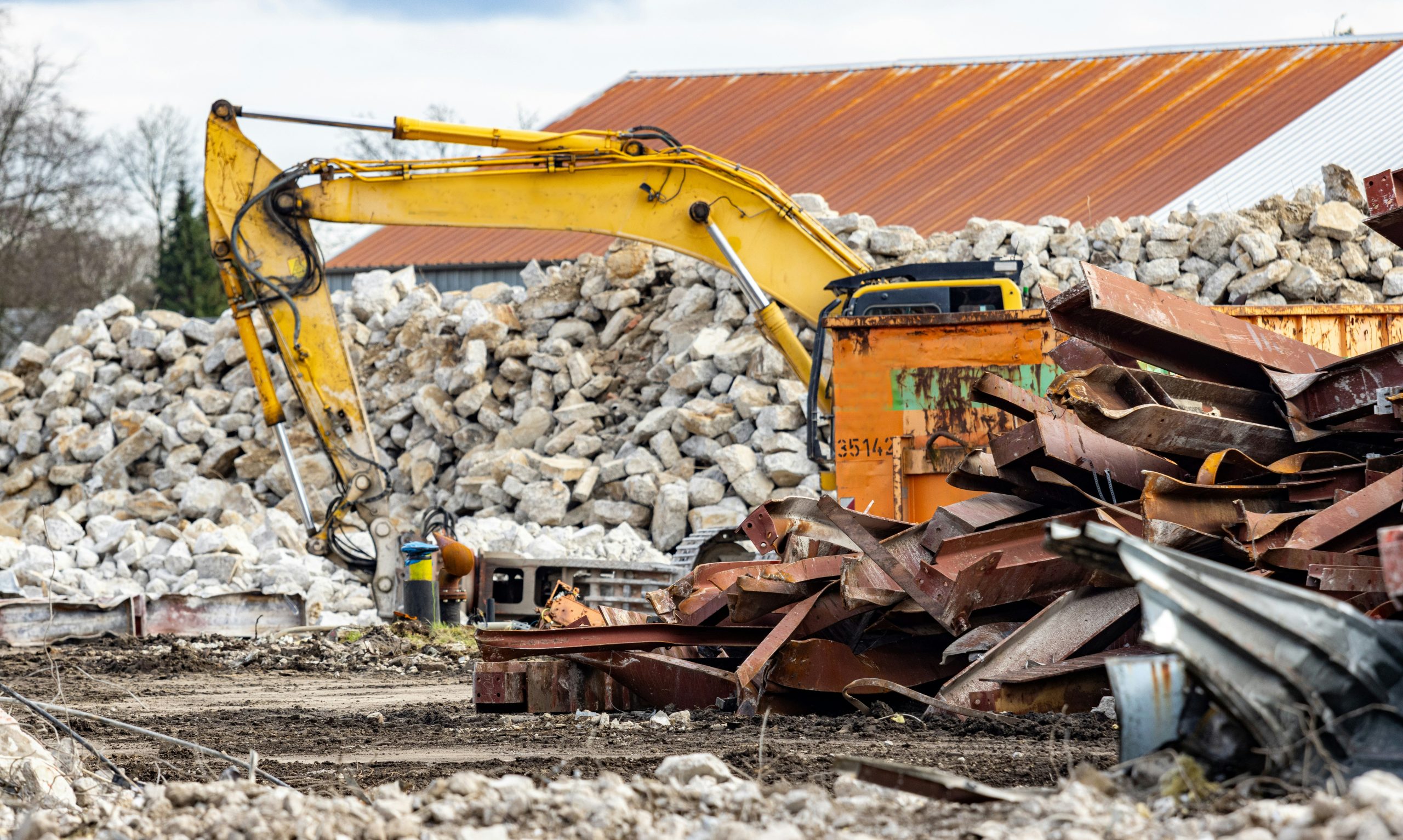 Excavator with hydraulic breaker beside a steel skip and piles of twisted girders and concrete rubble on a demolition jobsite